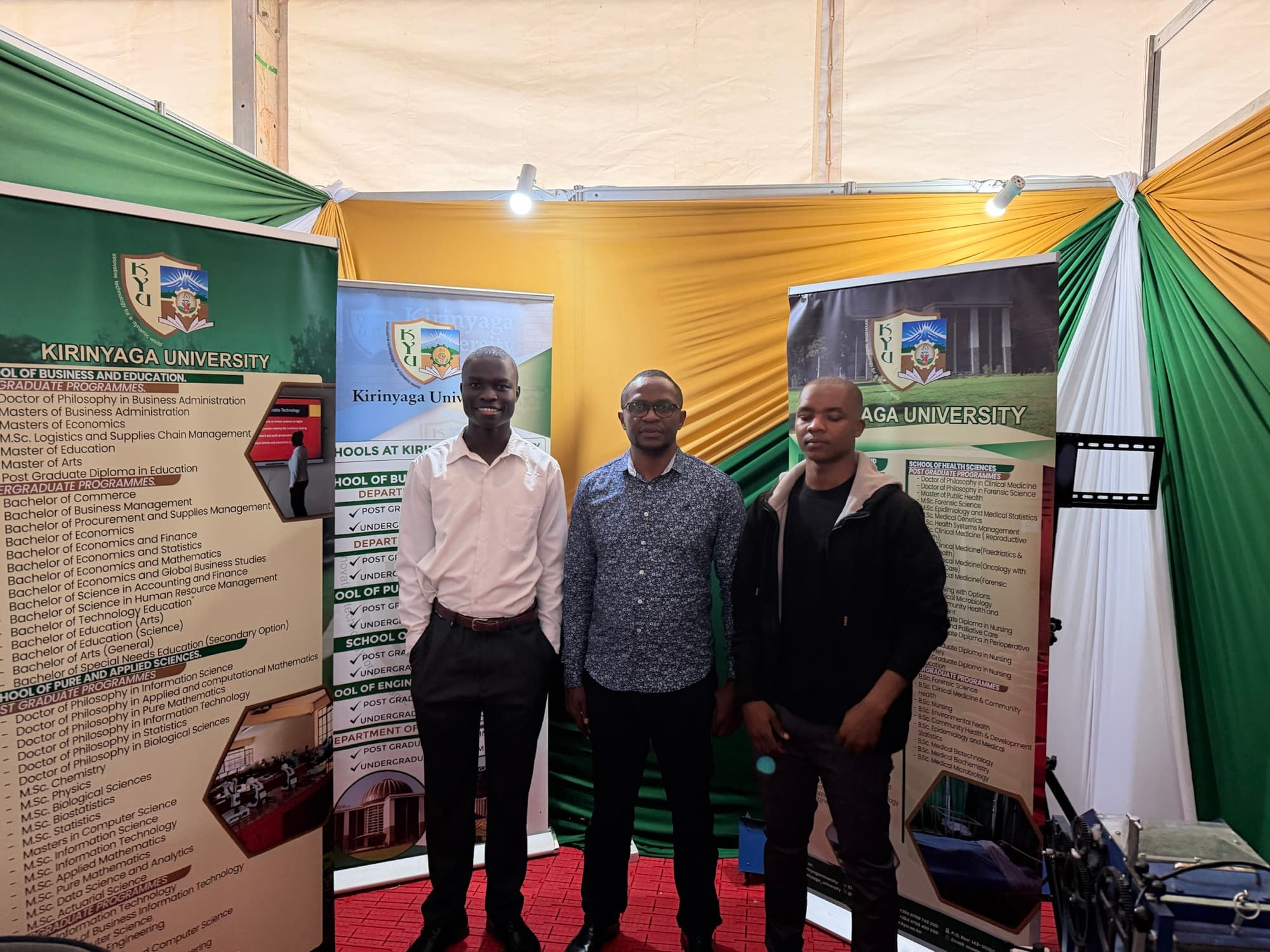 Hawona Rodgers, Dr. James Mwikya, and Mourice Oyugi inside the Kirinyaga University booth, posed in front of university programme banners.